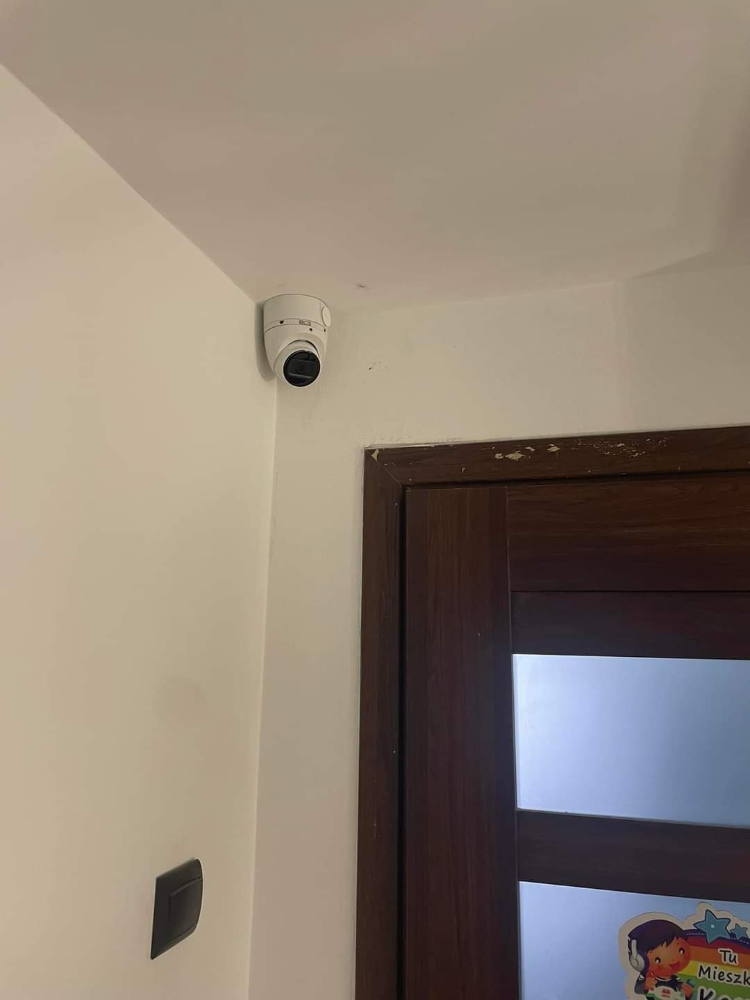 Biała kamera monitoringu kopułkowa zamontowana w rogu pokoju, widoczny włącznik światła i fragment brązowych drzwi z naklejką, na tle białej ściany i sufitu.