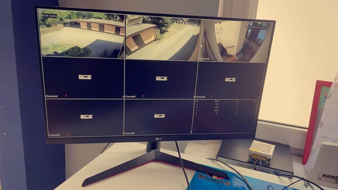 Monitor LCD prezentujący podgląd z kamer monitoringu BCS, z widokiem na posesję i otoczenie. Widoczne kanały i parametry transmisji. Sprzęt na biurku.