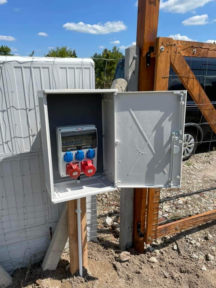Otwarta, szara skrzynka elektryczna z gniazdami (czerwone i niebieskie) zamontowana na drewnianym słupku, obok ogrodzenia i tymczasowego budynku. Słoneczny dzień.