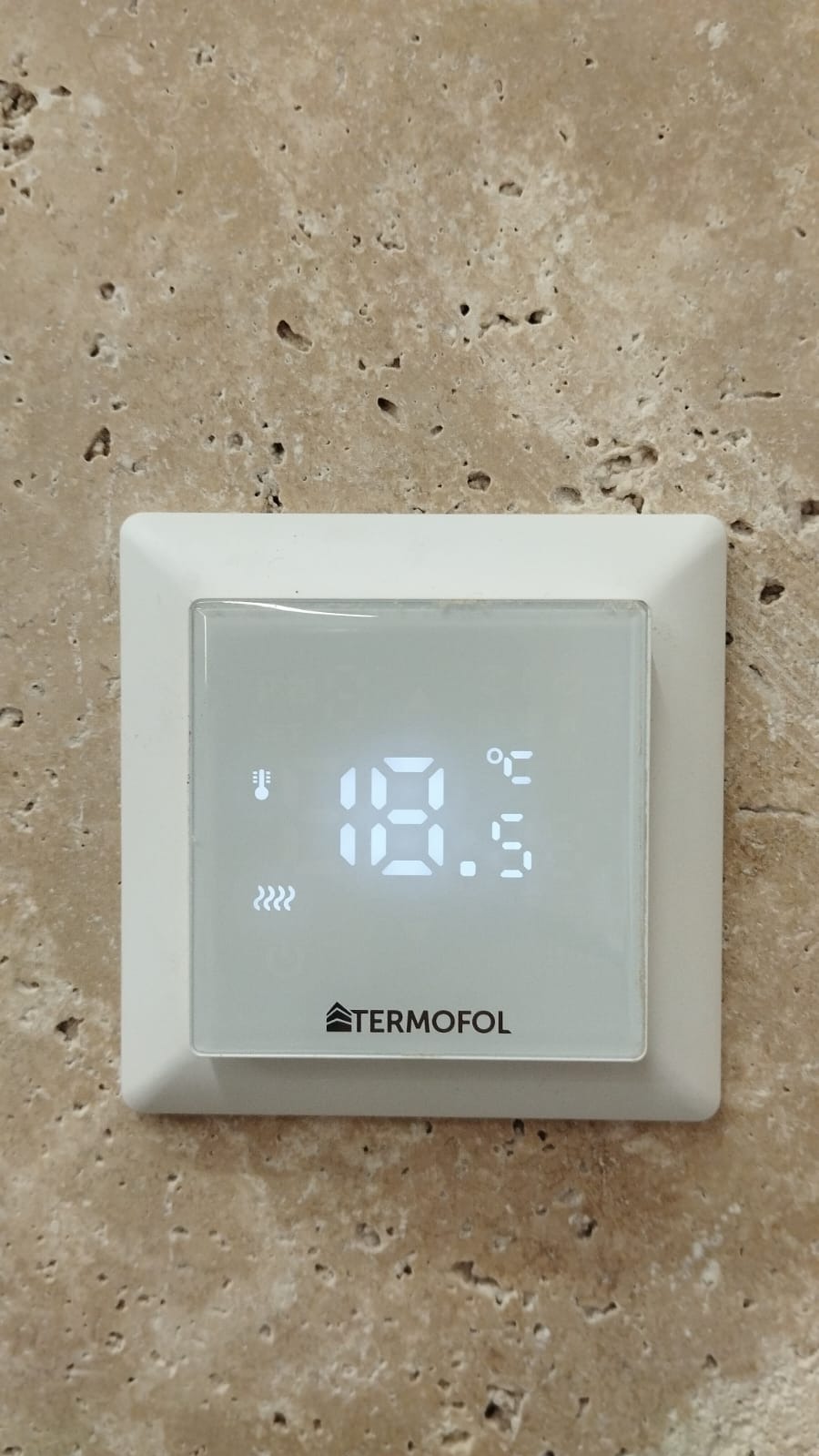 Biały, kwadratowy termostat marki Termofol zamontowany na beżowej ścianie z widoczną temperaturą 18.5 stopni Celsjusza, z bliska.