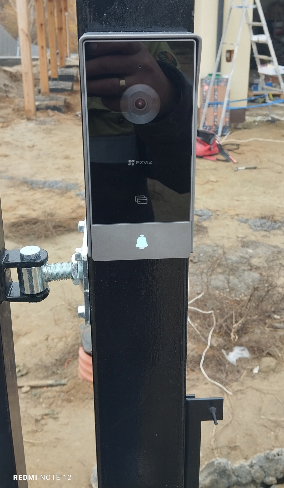 Zamontowany wideodomofon EZVIZ na czarnym słupku bramy, widoczny obiektyw kamery i czytnik kart. W tle elementy konstrukcyjne i drabina.