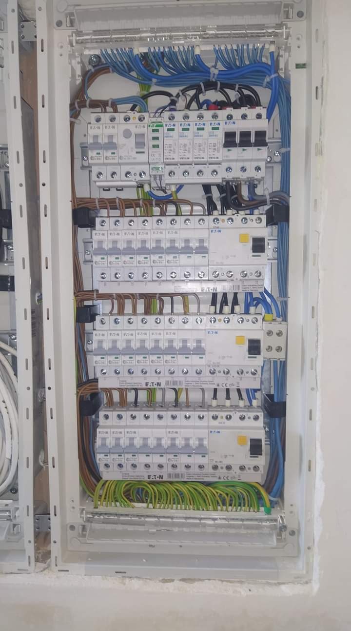 Rozdzielnia elektryczna z licznymi bezpiecznikami EATON, okablowaniem w kolorach: niebieskim, brązowym, zielono-żółtym. Widok z przodu na białą skrzynkę rozdzielczą w ścianie.