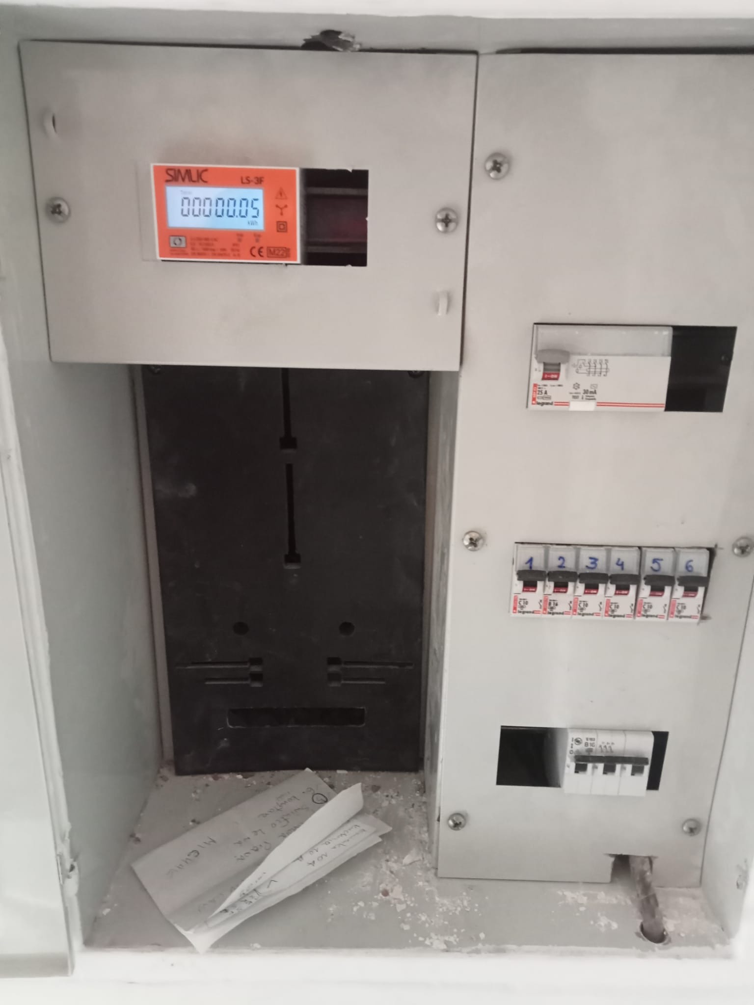 Szara skrzynka elektryczna z licznikiem SIMLIC LS-3F, bezpiecznikami i notatkami leżącymi na dnie. Widoczne ślady montażu.