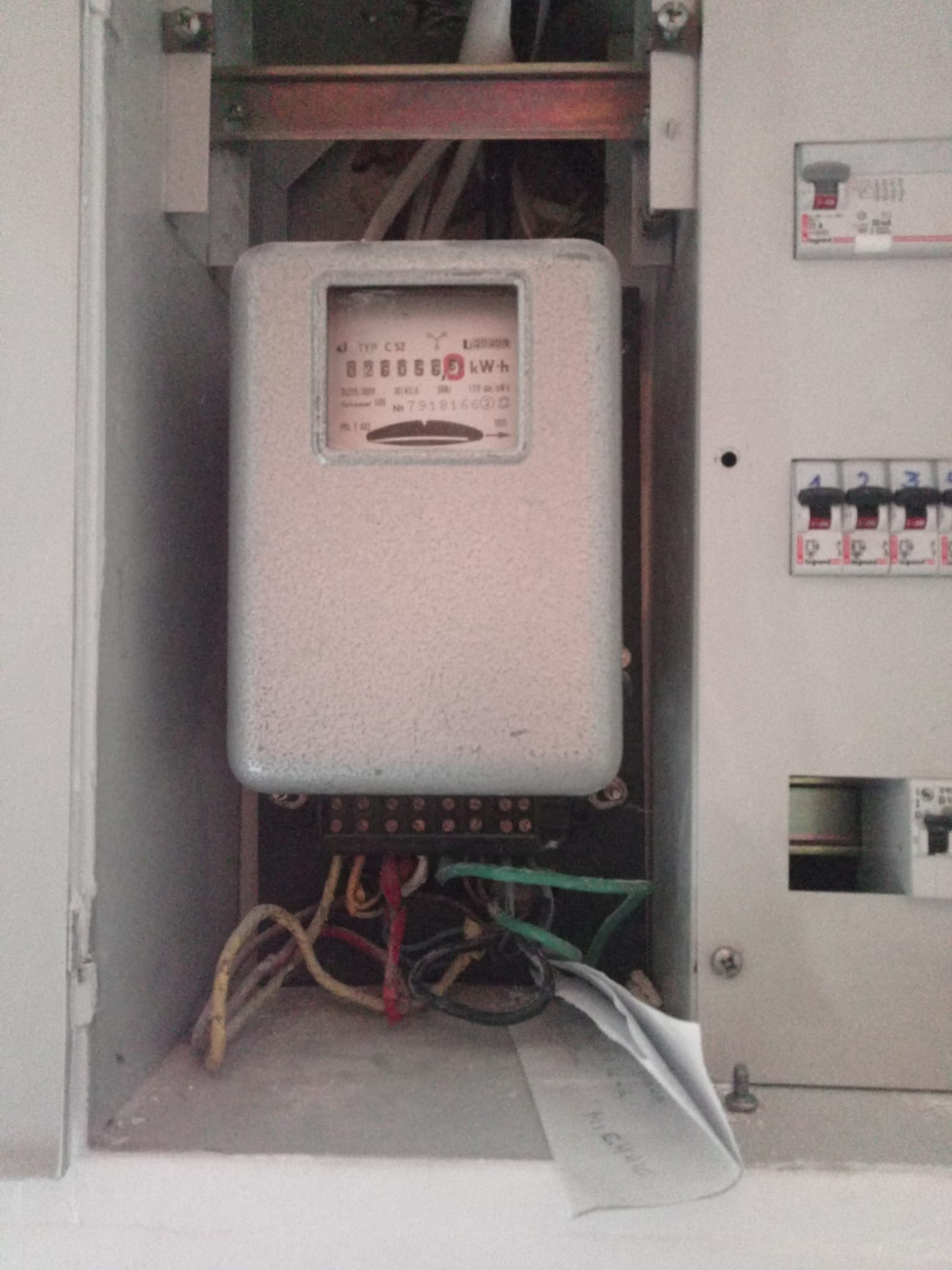 Szary licznik energii elektrycznej w skrzynce rozdzielczej, widoczne przewody i wyłączniki. Na dole kartka z notatkami.