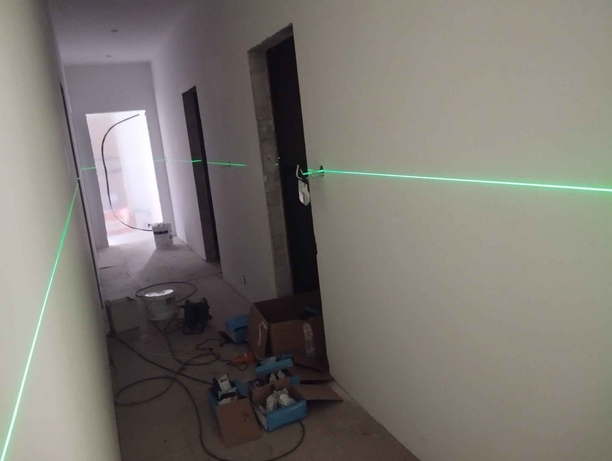 Wnętrze w trakcie remontu z użyciem lasera krzyżowego do wyznaczania linii na ścianach. Widoczne narzędzia i materiały budowlane. Oświetlenie robocze.