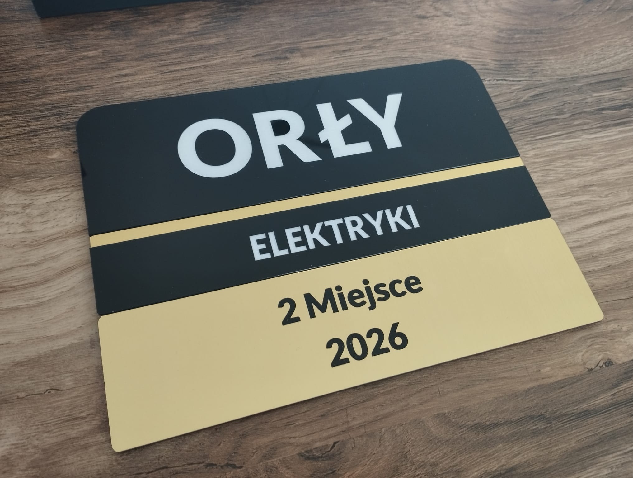 Tabliczka z napisem 'Orły Elektryki 2 Miejsce 2026' na drewnianym tle. Czarno-złota kolorystyka, nowoczesny design. Nagroda dla firmy elektrycznej.