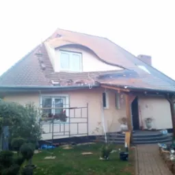 Dom z częściowo zdjętym pokryciem dachowym. Widoczne rusztowania i narzędzia sugerują trwający remont. Uwagę zwraca nietypowy kształt dachu z oknem w lukarnie.