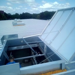 Daniel Brzózka - Metalowy świetlik dachowy otwarty na dachu z blachy trapezowej, widoczne uszczelnienie pianką montażową i fragment sylwetki osoby wewnątrz. Błękitne niebo w tle.