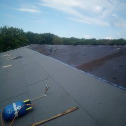 Daniel Brzózka - Układanie papy termozgrzewalnej na dachu z użyciem palnika i butli gazowej. Widoczne narzędzia pracy i fragment pokrycia dachowego na tle błękitnego nieba.