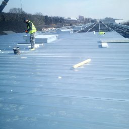 Daniel Brzózka - Pracownik w kamizelce na srebrnym dachu z blachy trapezowej montuje świetliki dachowe, w tle konstrukcja dachu i dźwig budowlany.