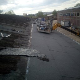 Daniel Brzózka - Fragment dachu z widocznym etapem prac: stara, zniszczona powierzchnia obok nowej, pokrytej papą. Dźwig z platformą roboczą w tle, budynek w oddali.