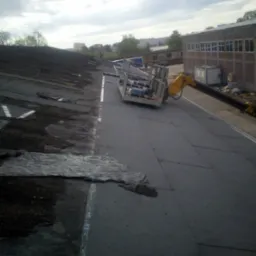 Fragment dachu z widocznym etapem prac: stara, zniszczona powierzchnia obok nowej, pokrytej papą. Dźwig z platformą roboczą w tle, budynek w oddali.