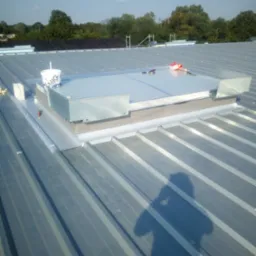 Montaż okna dachowego na blaszanym dachu, widoczne narzędzia i puszka farby. W tle zieleń drzew i błękit nieba, cień osoby robiącej zdjęcie.