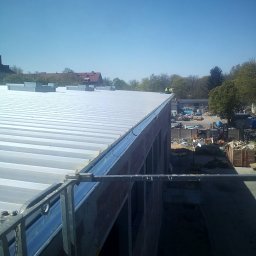 Daniel Brzózka - Montaż blachy trapezowej na dachu budynku w trakcie budowy. Widoczne rynny i elementy konstrukcyjne. W tle drzewa i budynki.