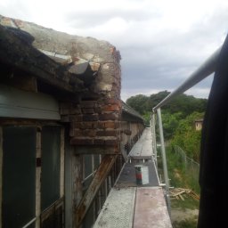 Daniel Brzózka - Zniszczony fragment dachu z ceglaną ścianą i blachą, widoczny z rusztowania na tle zachmurzonego nieba i zieleni. Prace remontowe w toku, z puszką farby na podeście.