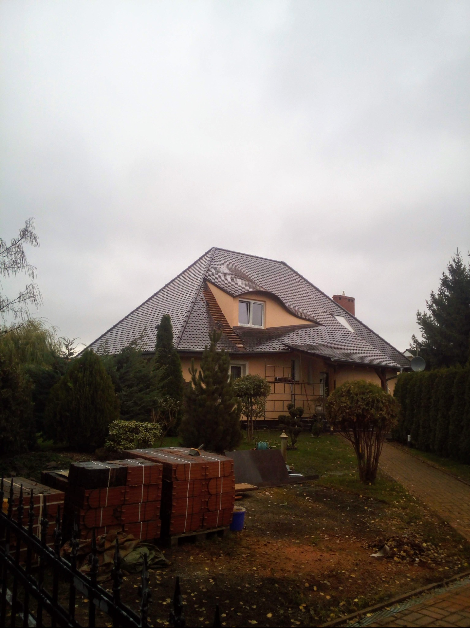 Dom z ciemną dachówką w pochmurny dzień. Na pierwszym planie palety z cegłami. Widoczny komin i okno dachowe. Otoczenie domu to drzewa iglaste i ścieżka.
