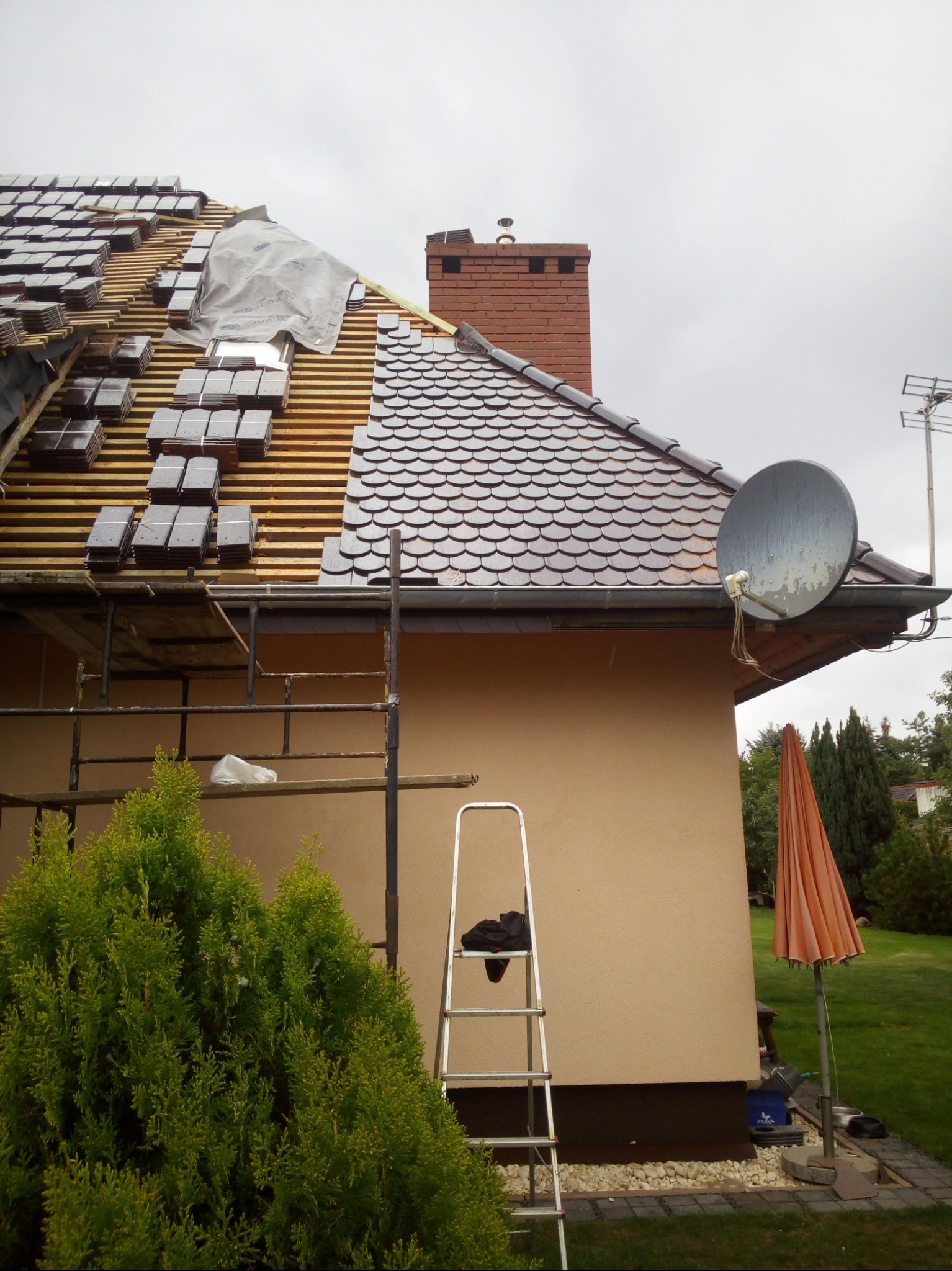Montaż dachówki karpiówki na drewnianej konstrukcji dachu, widoczny komin, rusztowanie i drabina oparta o ścianę budynku. Część dachu przykryta folią.