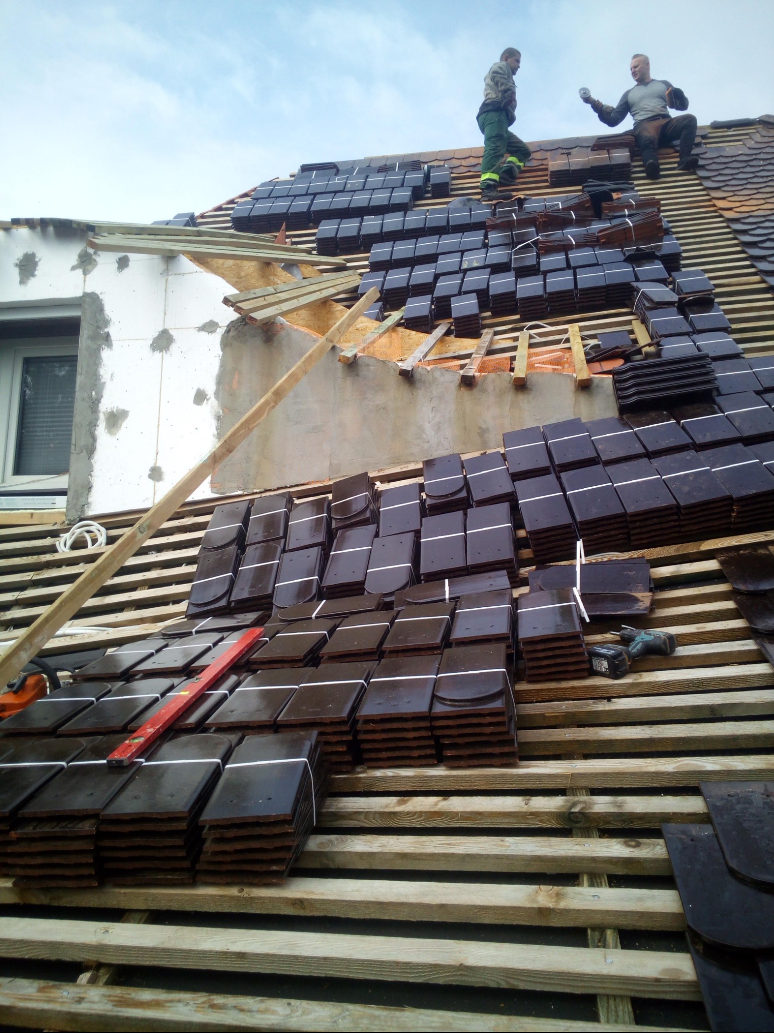 Prace dekarskie w toku: ułożone dachówki w regularnych rzędach na drewnianej konstrukcji dachu. Dwóch dekarzy na szczycie dachu, jeden siedzi, drugi stoi.