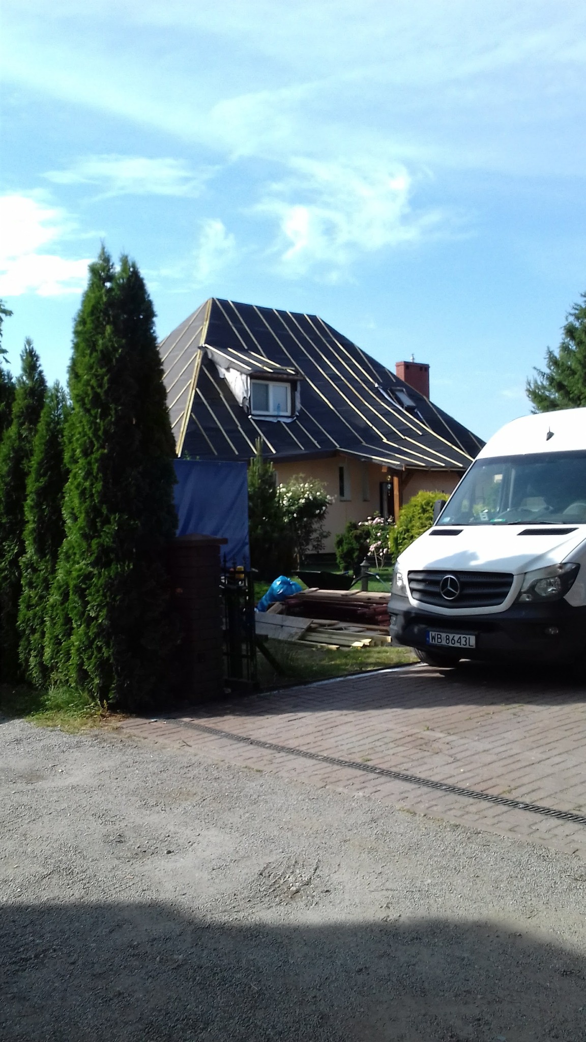 Dom jednorodzinny z dachem w trakcie krycia, widoczne łaty i membrana. Na pierwszym planie zaparkowany biały van i fragment podwórka z zielenią.