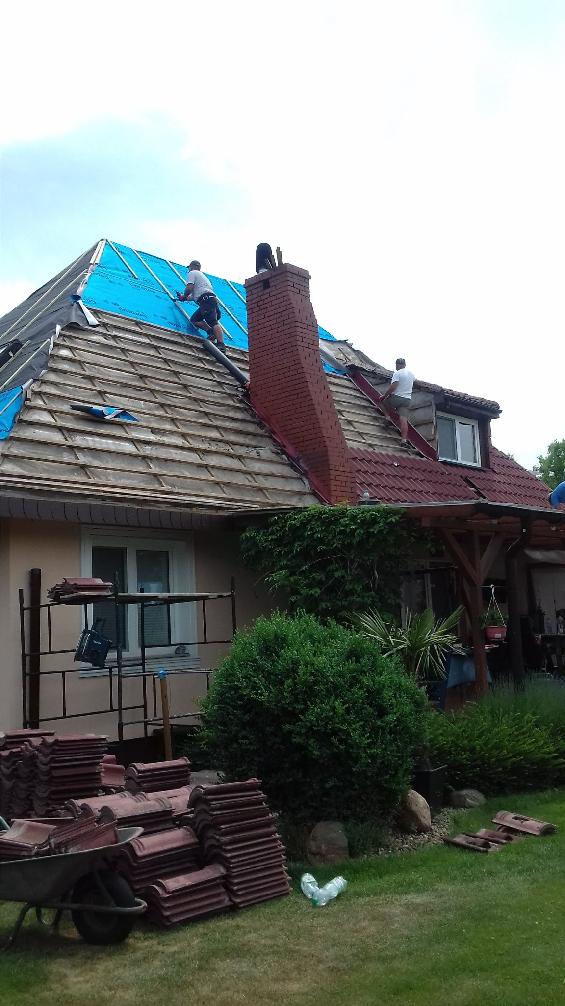 Prace dekarskie: robotnicy na dachu krytym częściowo papą i dachówką, z kominem z czerwonej cegły. Na trawniku sterta dachówek i taczka.