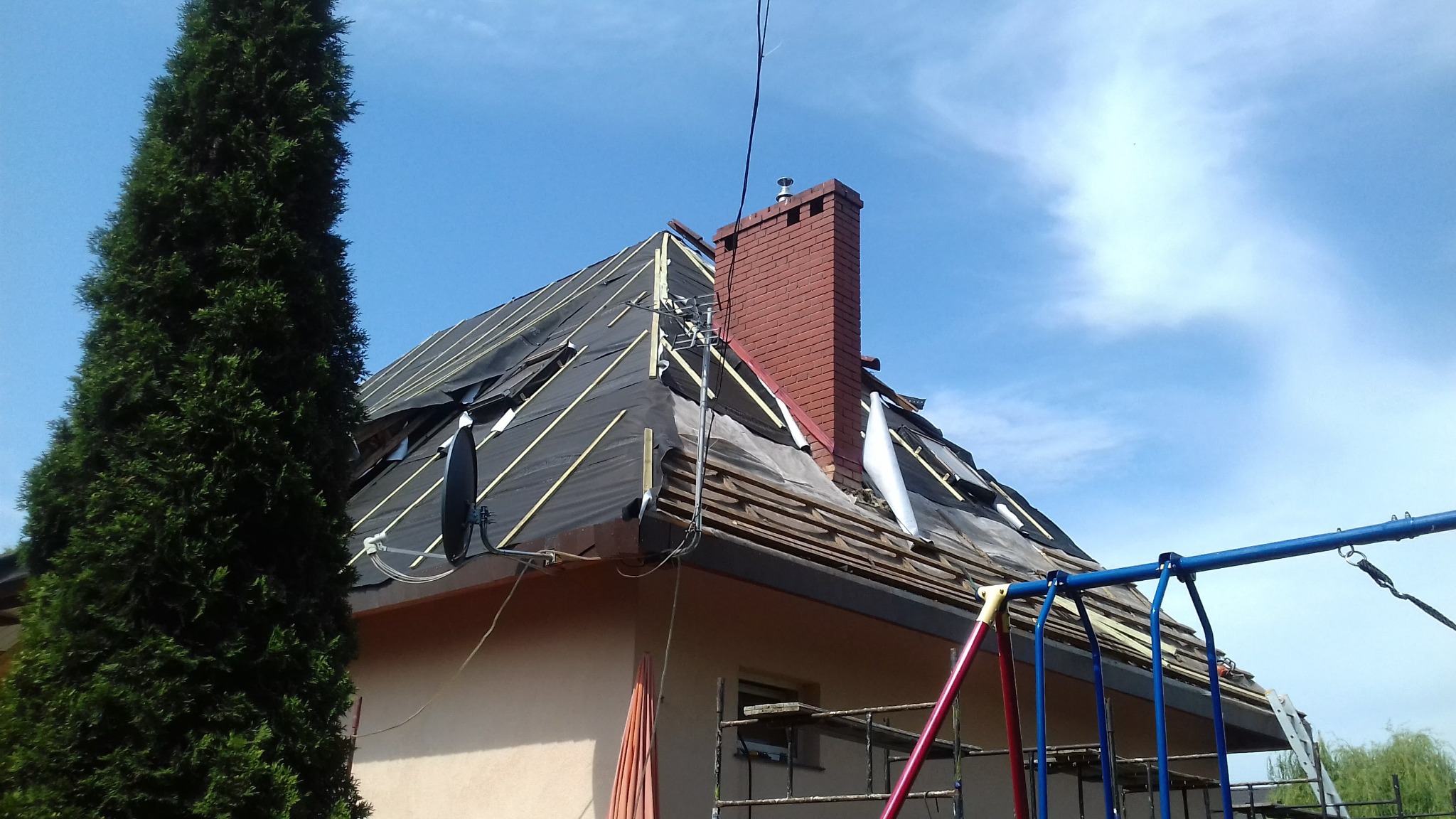 Dom w trakcie wymiany pokrycia dachowego, widoczna czarna membrana i fragmenty starego poszycia. Czerwony komin i drabina przy ścianie.