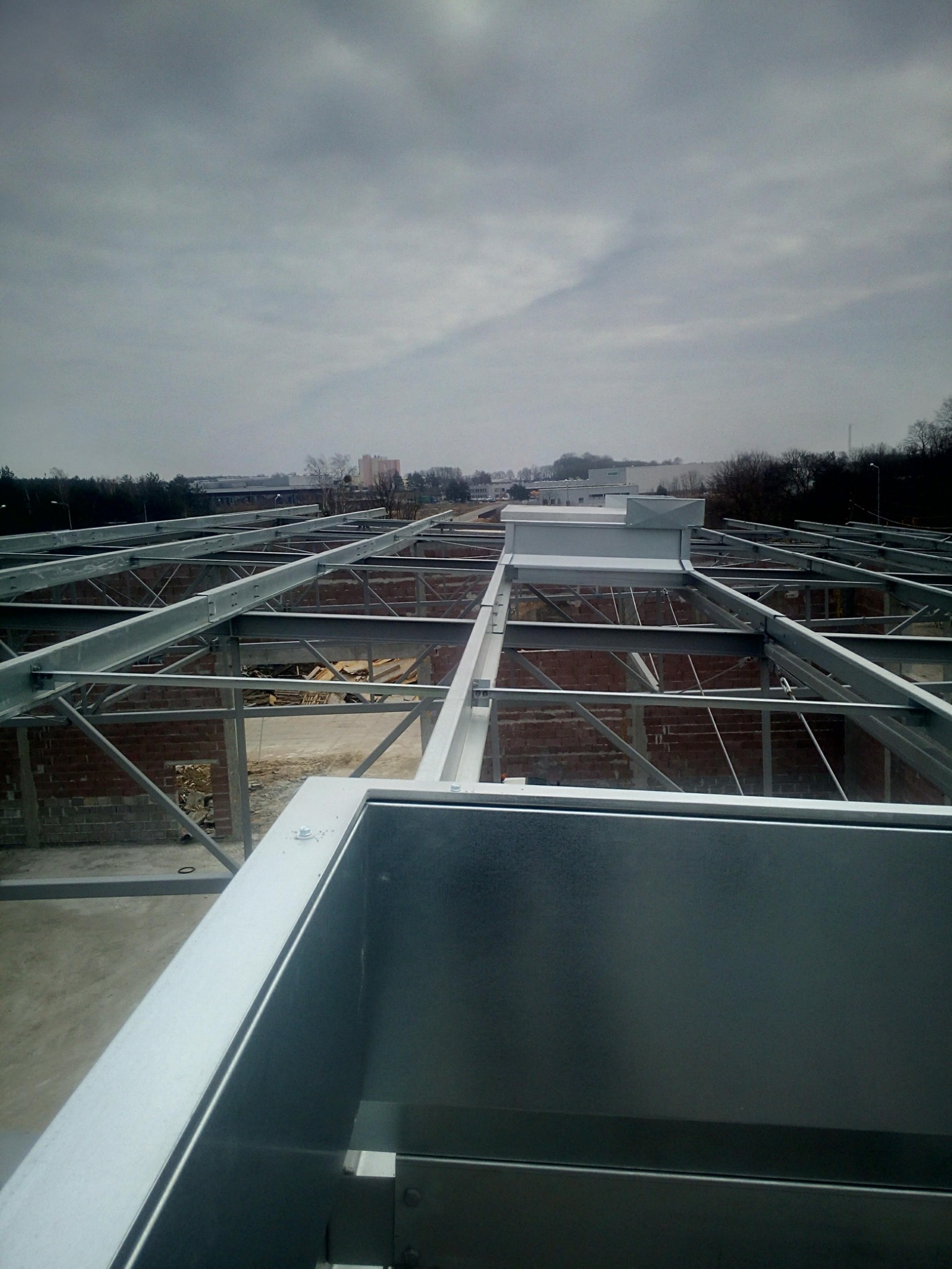 Stalowa konstrukcja dachu budynku w trakcie budowy, widok z góry, z elementami murowanymi w tle i pochmurnym niebem.