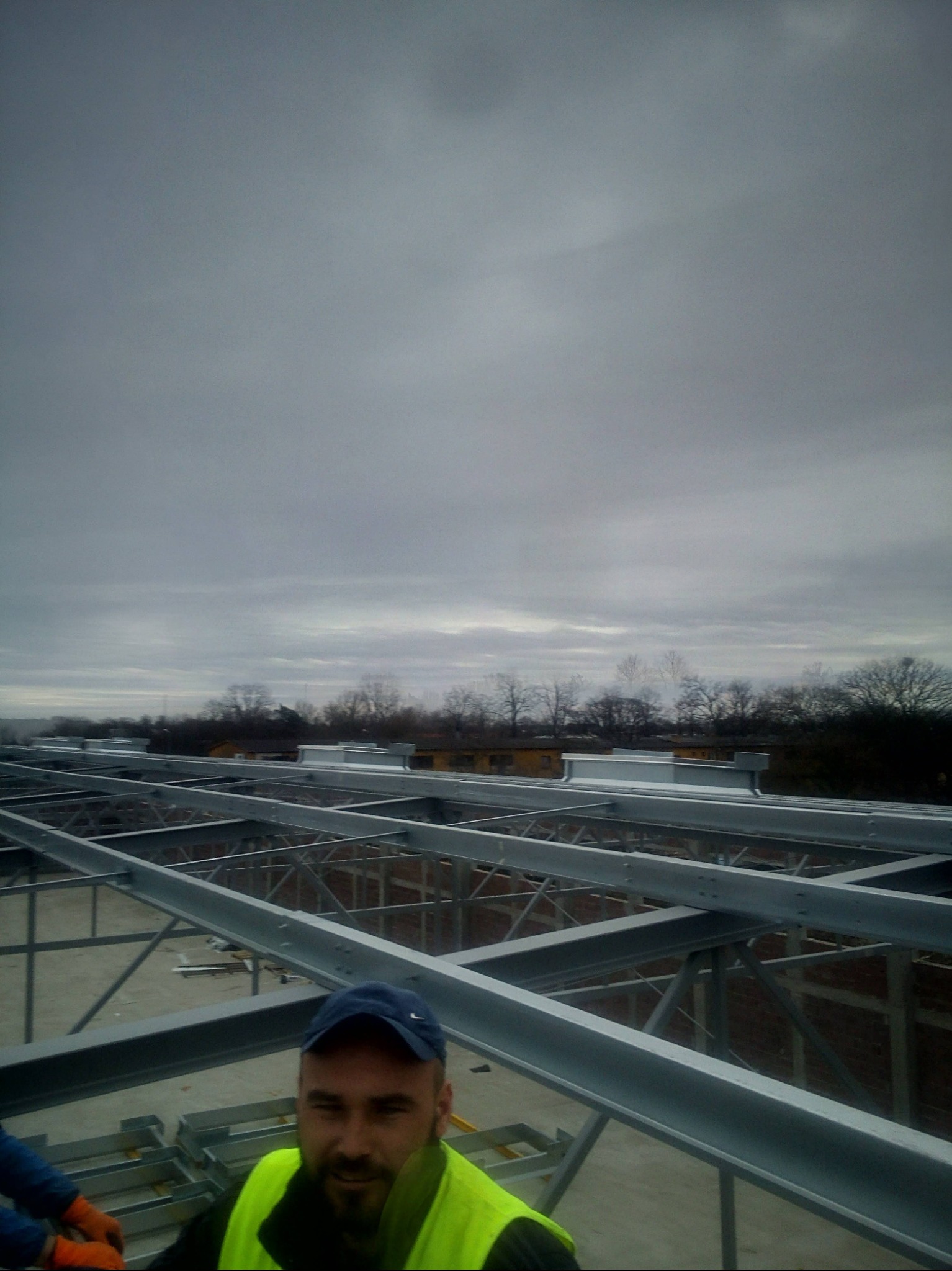 Stalowa konstrukcja dachu budynku w trakcie montażu, widok z góry. Pracownik w kamizelce odblaskowej i czapce w dolnej części kadru. Pochmurne niebo w tle.