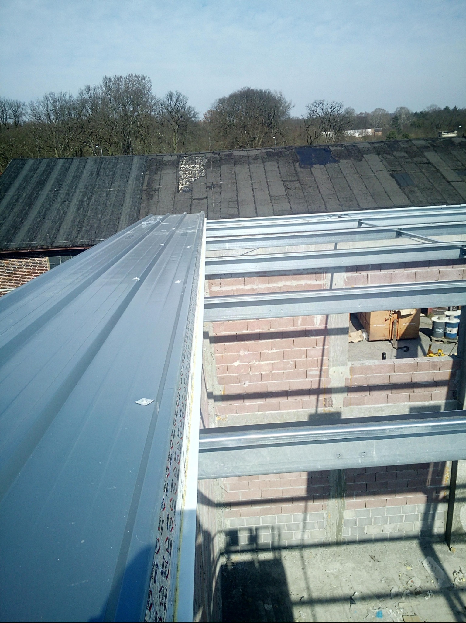 Nowa blacha trapezowa na dachu budynku w trakcie budowy, widok z góry na konstrukcję i częściowo zakryty stary dach w tle.