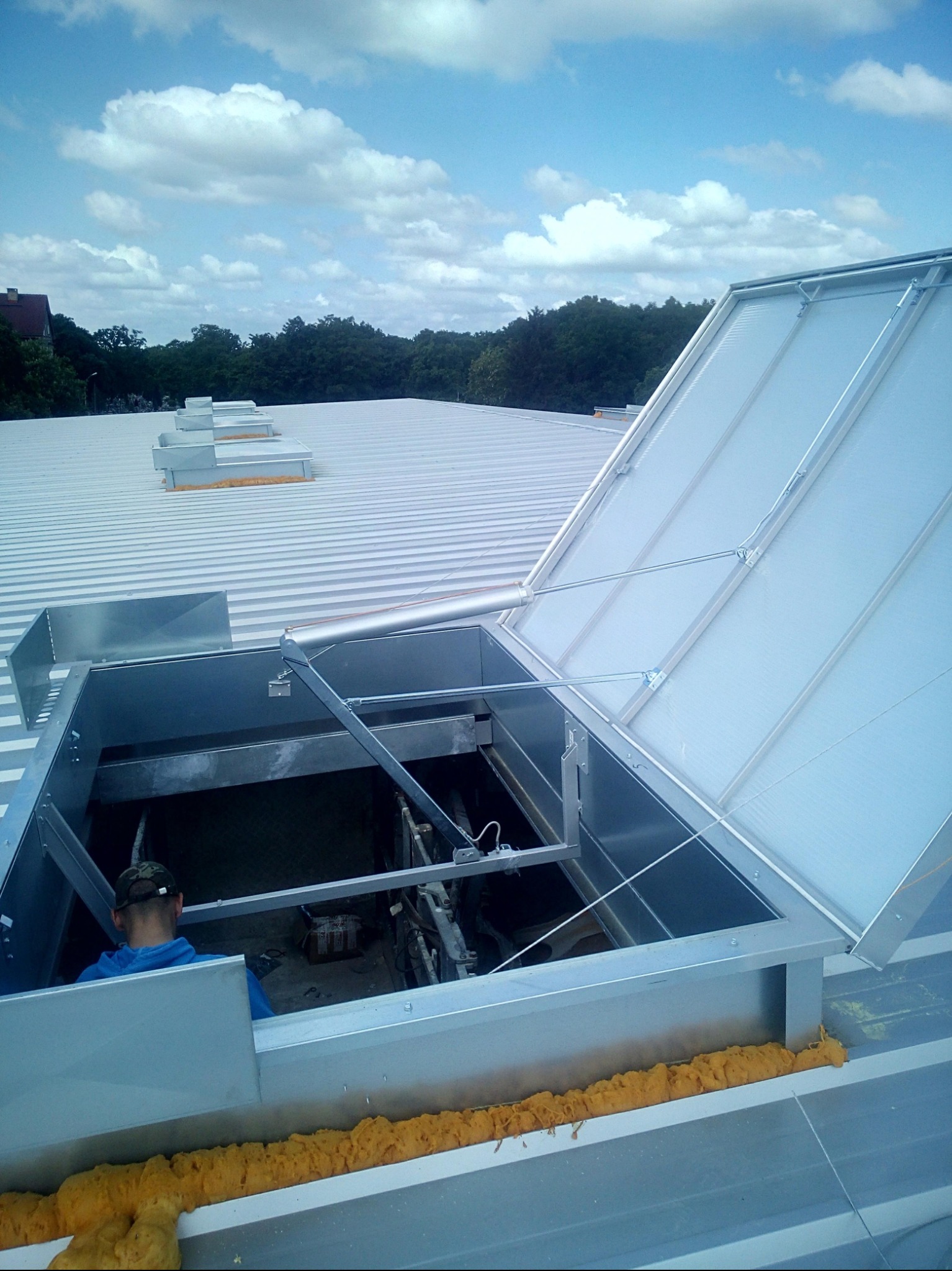 Metalowy świetlik dachowy otwarty na dachu z blachy trapezowej, widoczne uszczelnienie pianką montażową i fragment sylwetki osoby wewnątrz. Błękitne niebo w tle.