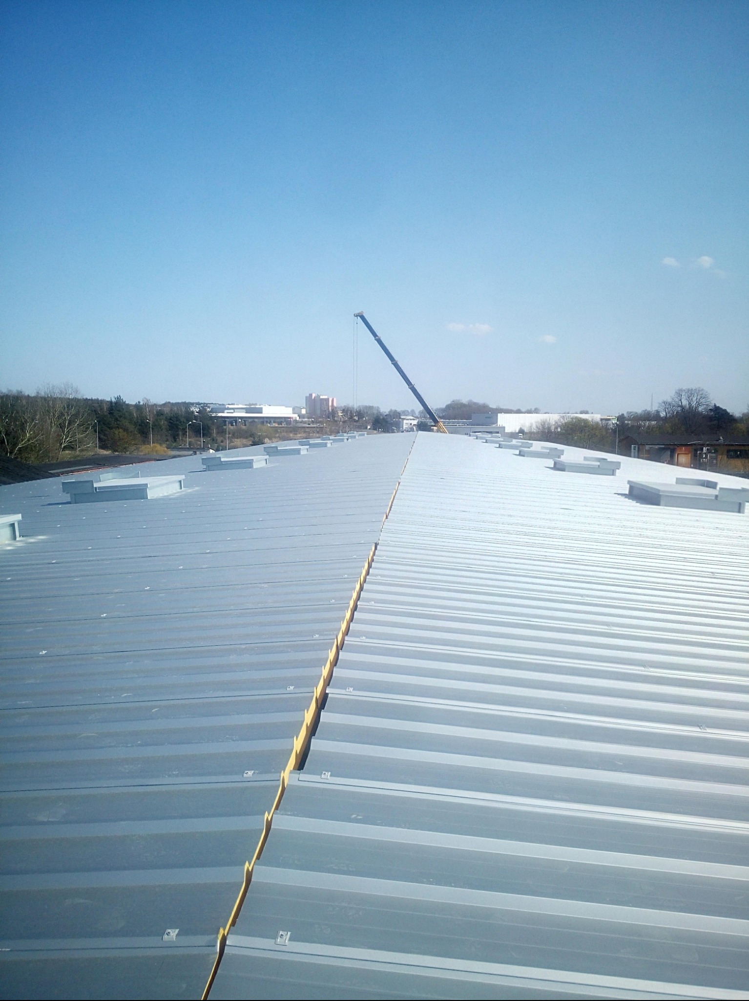 Długi, srebrny dach przemysłowy z licznymi świetlikami i dźwigiem w tle pod błękitnym niebem. Widok z góry, ukazujący konstrukcję dachu.