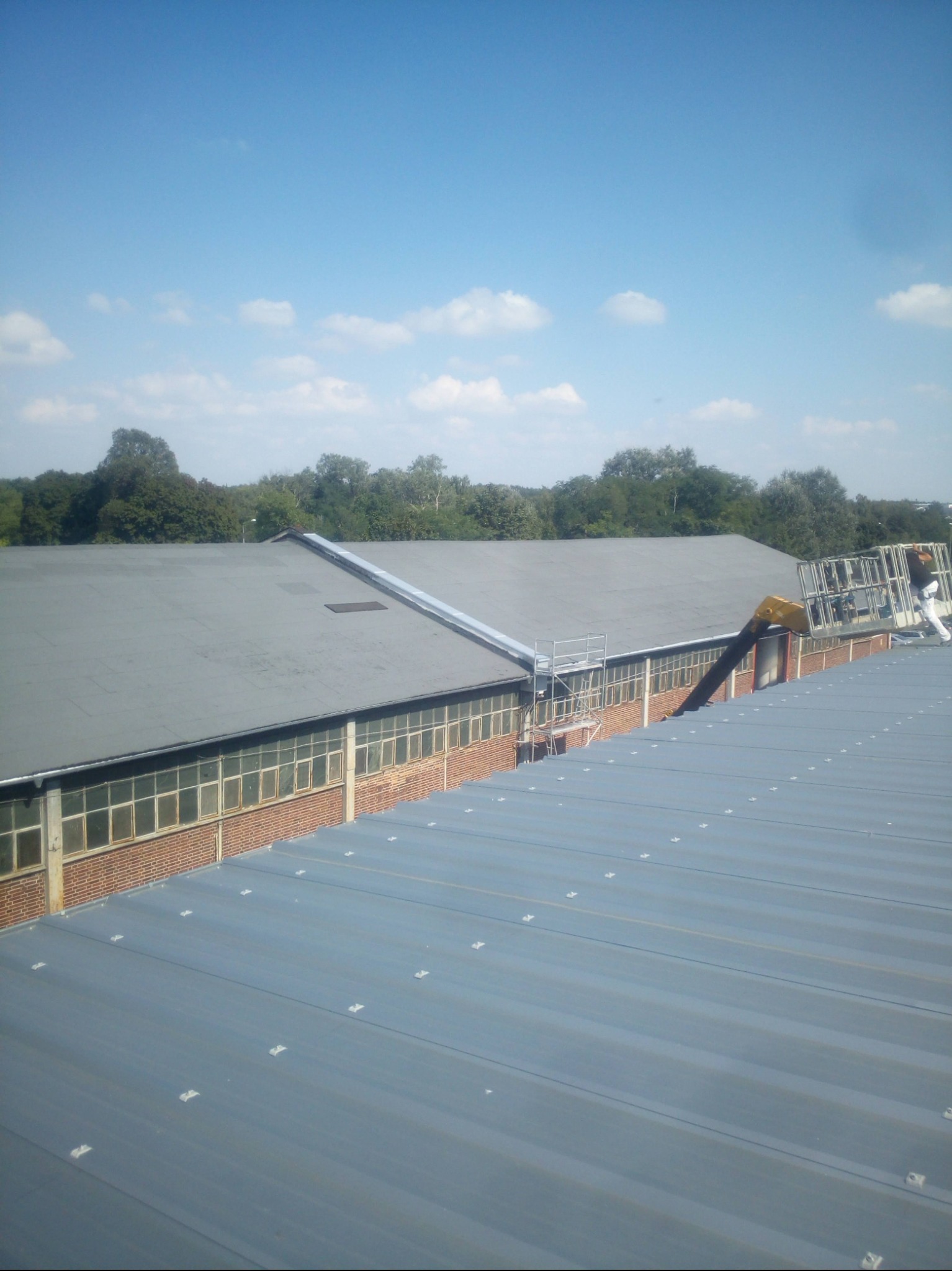 Szary dach z blachy trapezowej z widocznymi mocowaniami, w tle budynek z cegły i okien oraz zieleń drzew. Pracownik na podnośniku koszowym wykonuje prace na dachu.