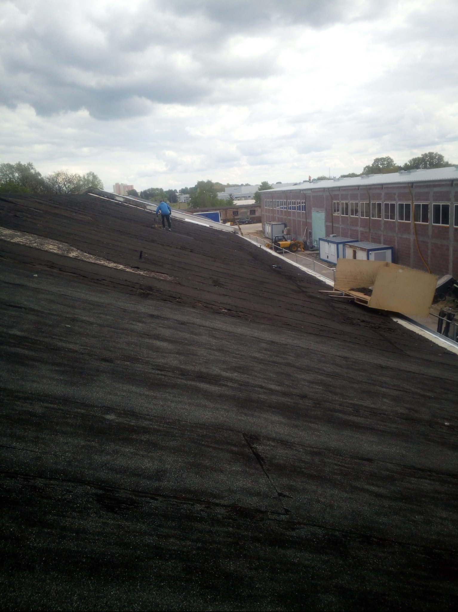 Praca na dachu budynku przemysłowego. Widoczny pracownik w niebieskiej kurtce na tle pochmurnego nieba i zabudowań. Fragment dachu z bliska, w tle budynek z licznymi oknami.