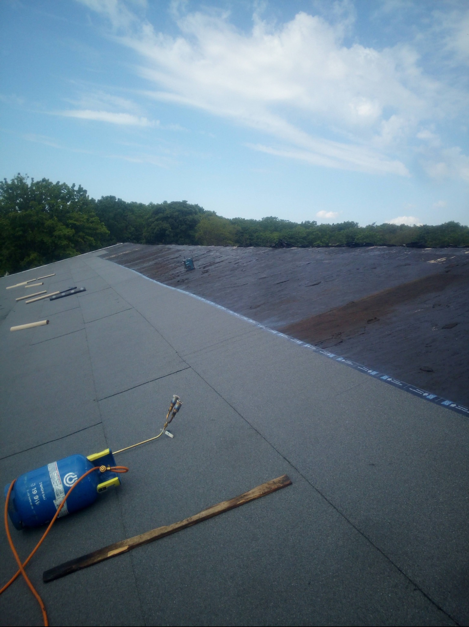 Układanie papy termozgrzewalnej na dachu z użyciem palnika i butli gazowej. Widoczne narzędzia pracy i fragment pokrycia dachowego na tle błękitnego nieba.