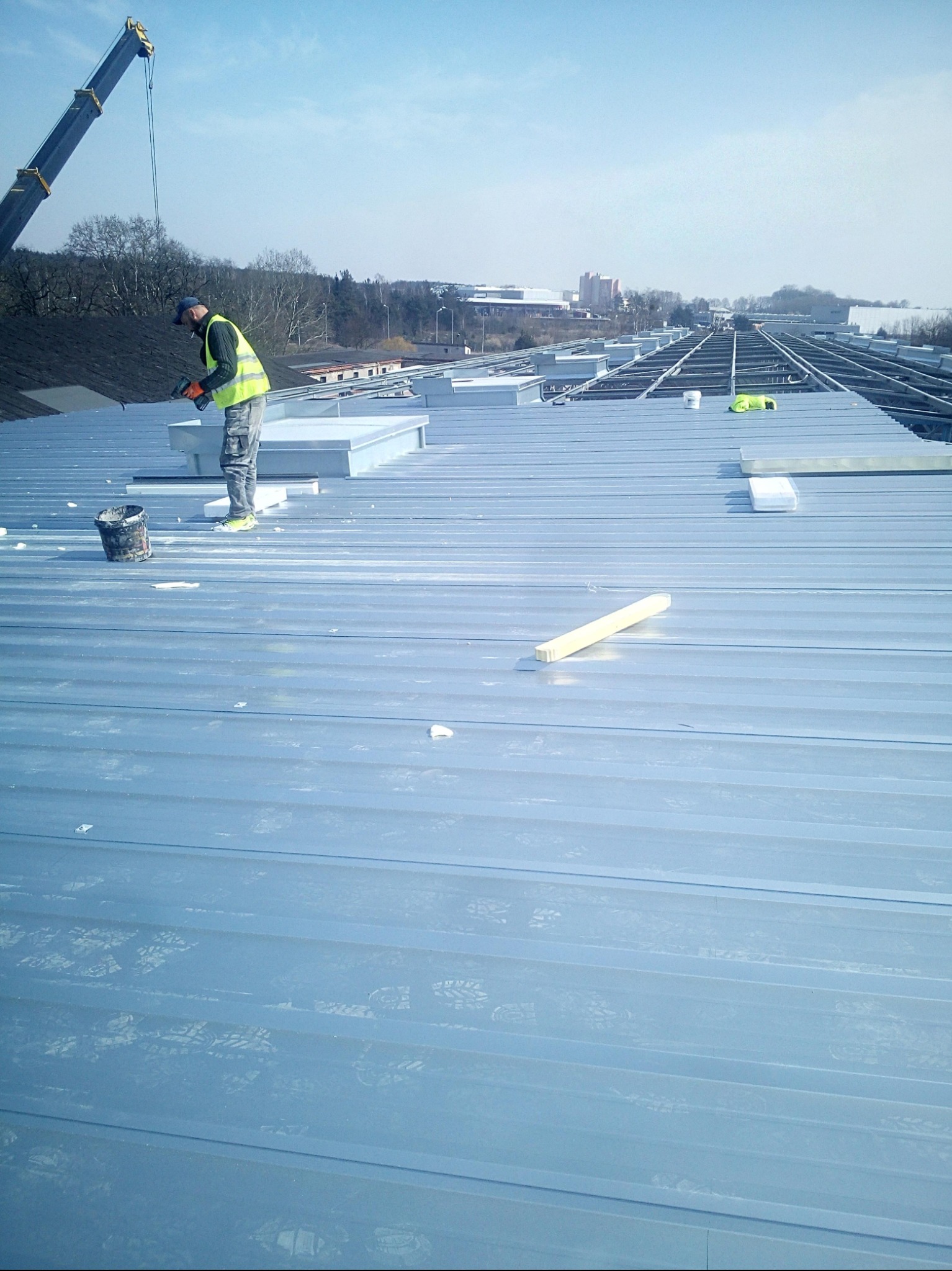 Pracownik w kamizelce na srebrnym dachu z blachy trapezowej montuje świetliki dachowe, w tle konstrukcja dachu i dźwig budowlany.