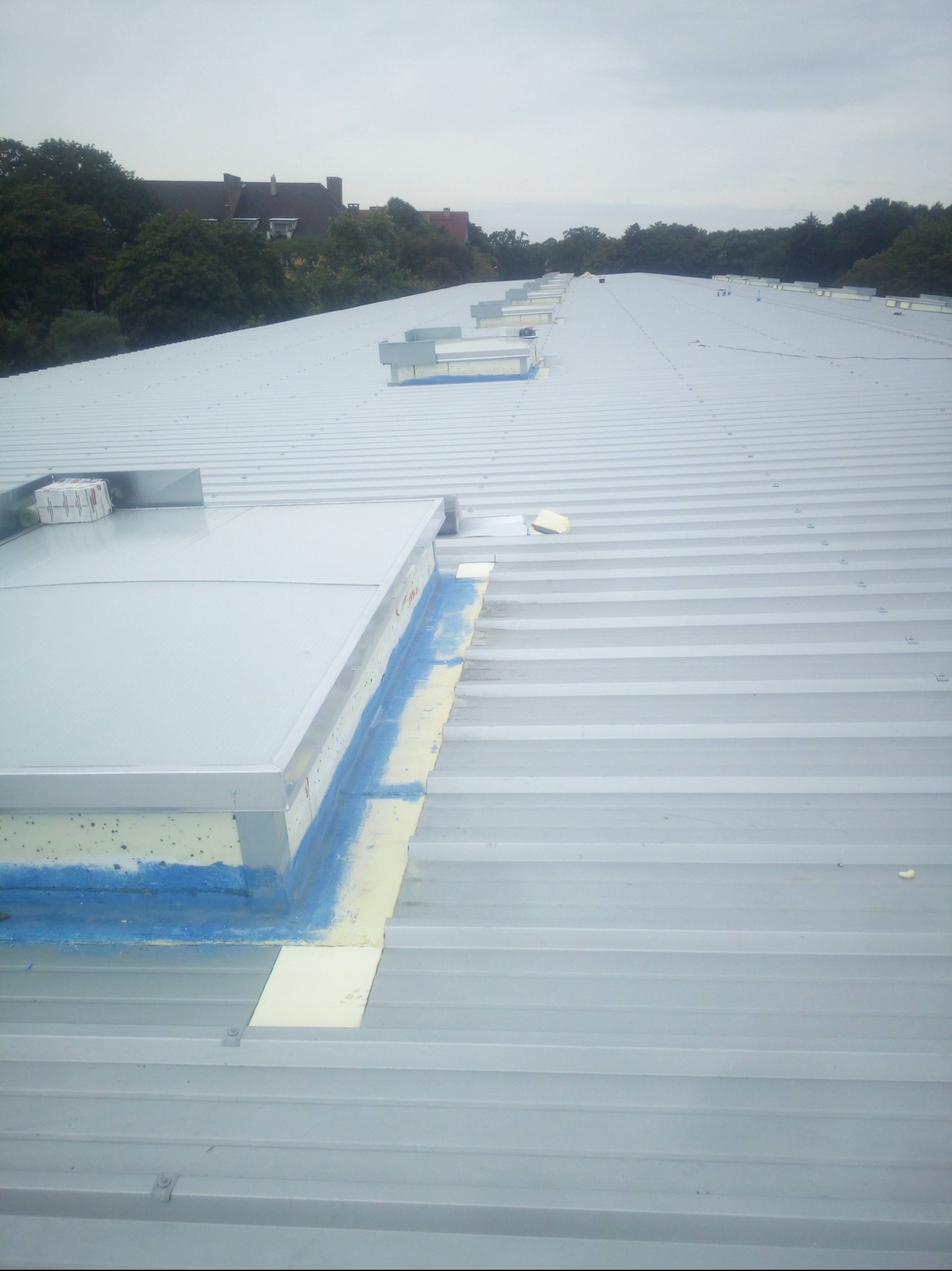 Szary, falisty dach przemysłowy z oknami dachowymi, uszczelnieniami i widocznymi mocowaniami, na tle zieleni i zabudowań w oddali.