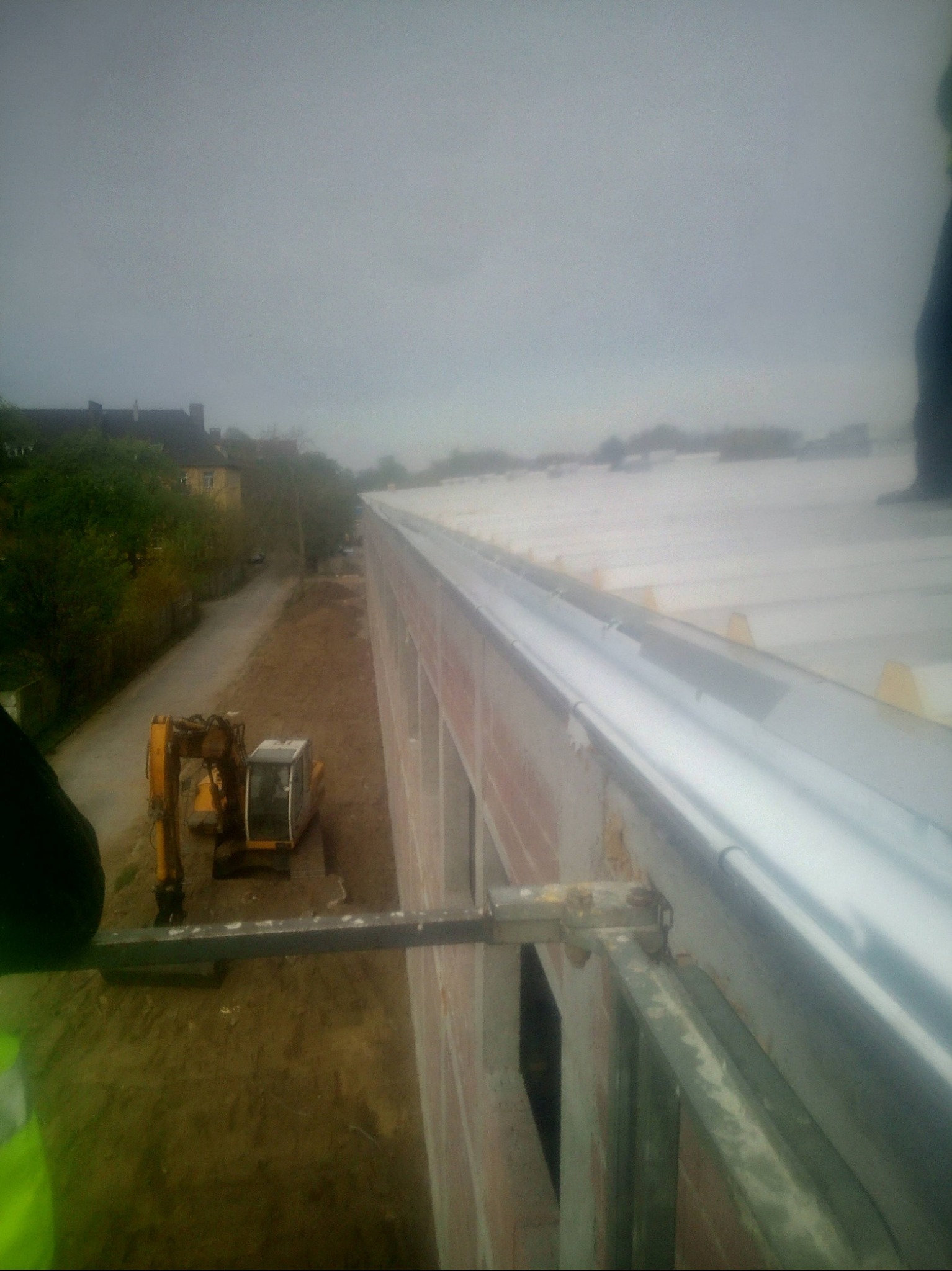 Widok z góry na długi, biały dach z zamontowanymi rynnami, obok budynku stoi koparka, pochmurne niebo w tle. Prace dekarskie w toku.