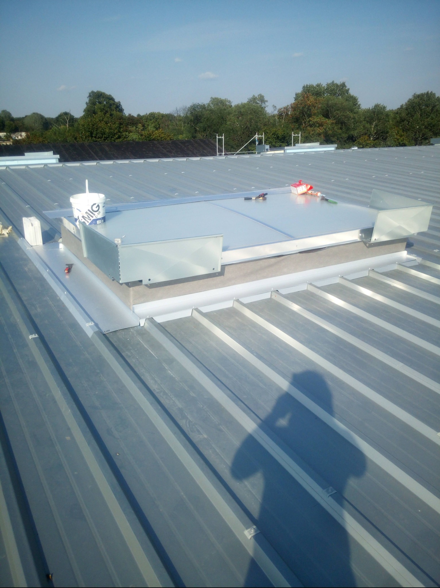 Montaż okna dachowego na blaszanym dachu, widoczne narzędzia i puszka farby. W tle zieleń drzew i błękit nieba, cień osoby robiącej zdjęcie.