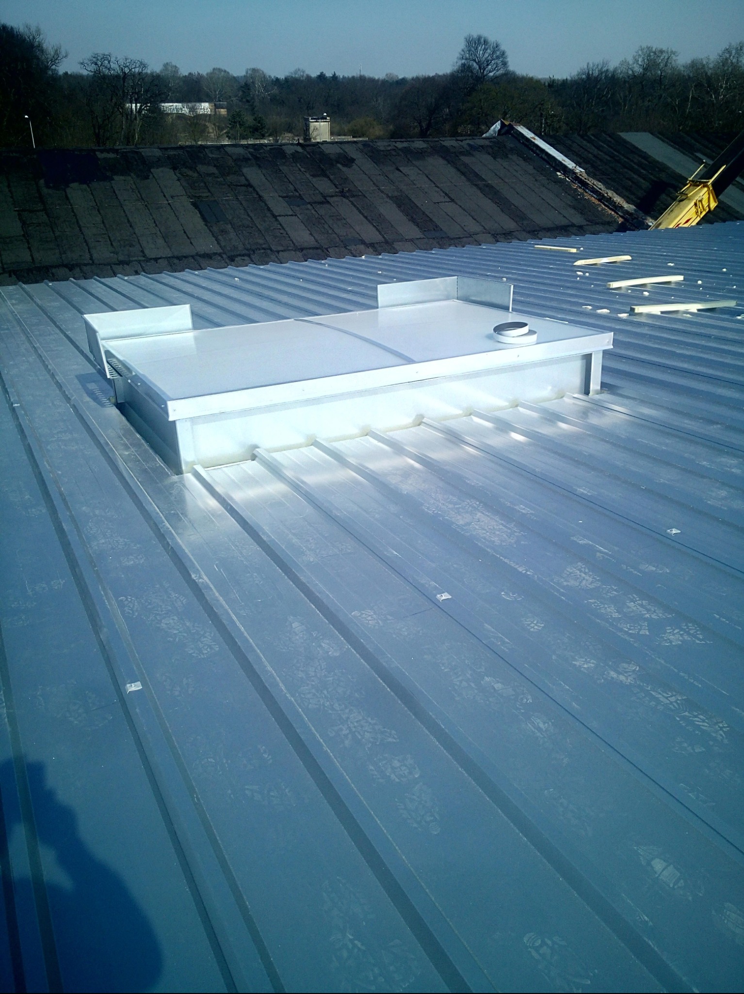 Blaszany dach z świetlikiem dachowym w kształcie prostopadłościanu. W tle widać budynki i drzewa. Dach z blachy trapezowej w kolorze srebrnym.