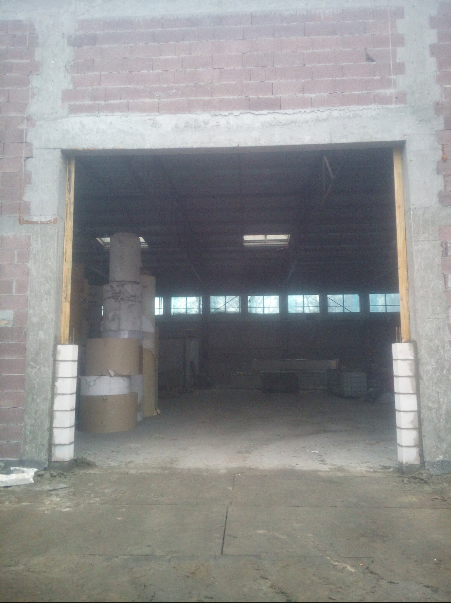 Wnętrze magazynu z widoczną konstrukcją dachu i oknami. Ceglany mur z betonowymi elementami wykończeniowymi tworzy obramowanie otworu wjazdowego.