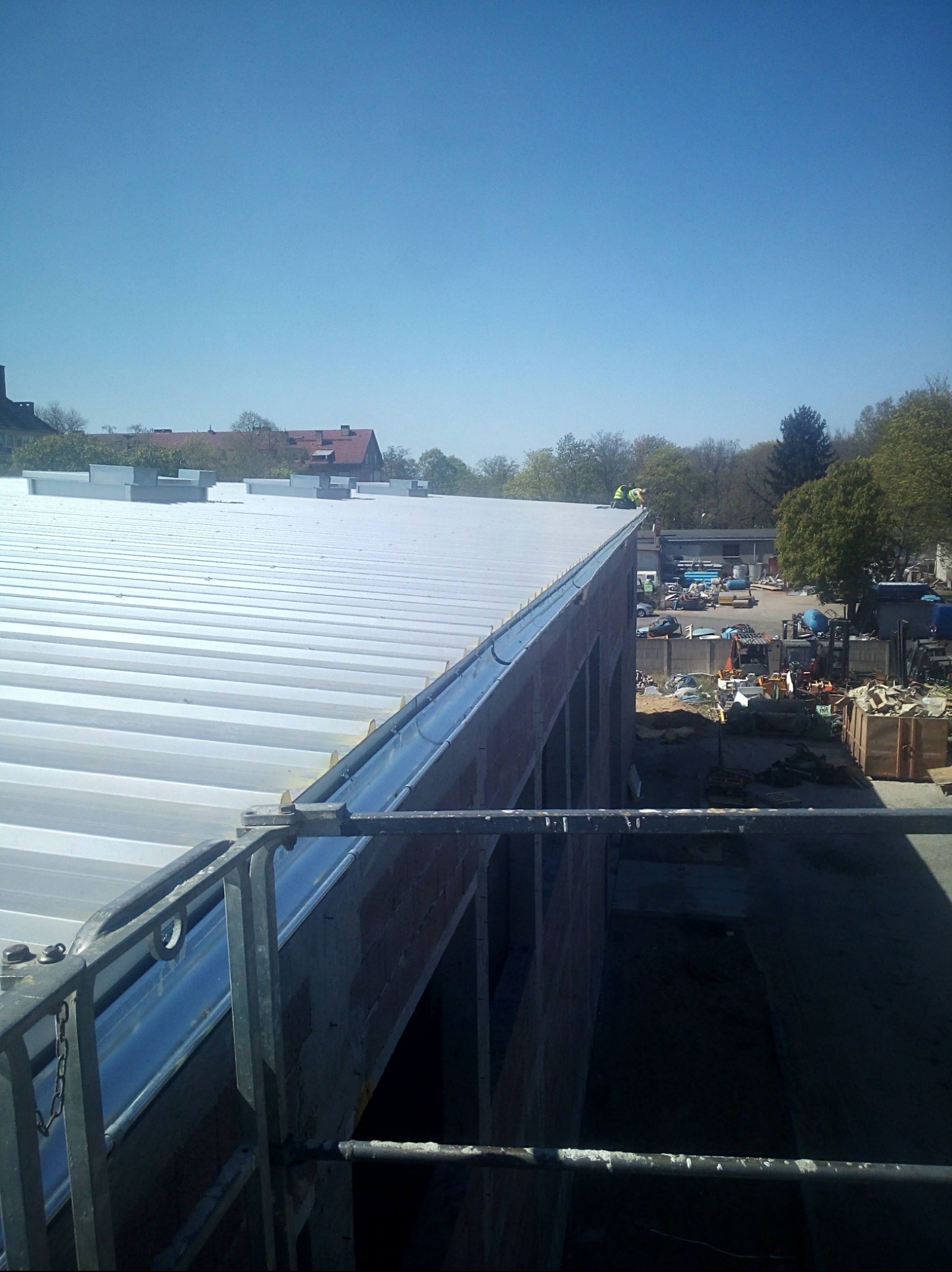 Montaż blachy trapezowej na dachu budynku w trakcie budowy. Widoczne rynny i elementy konstrukcyjne. W tle drzewa i budynki.