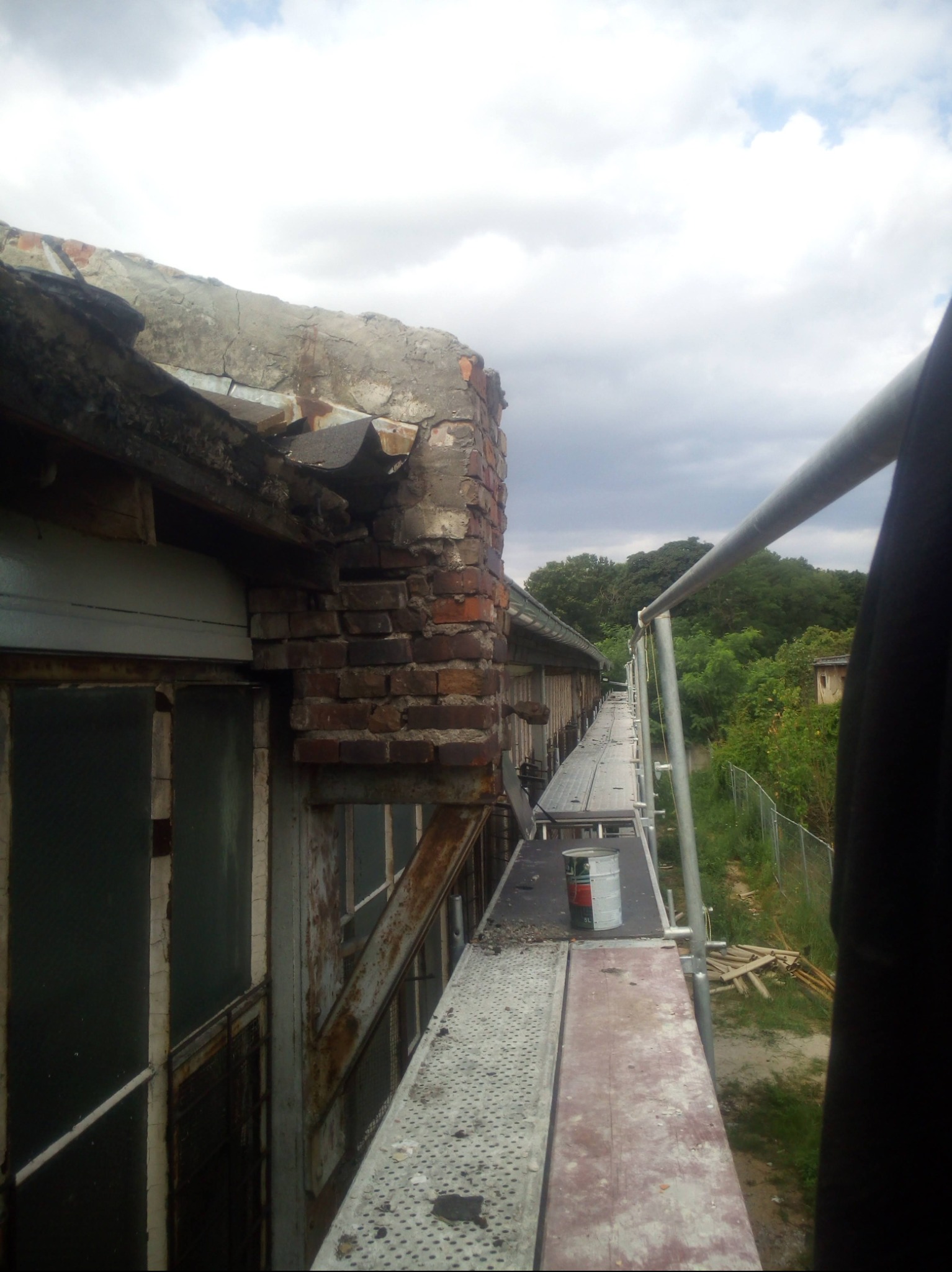 Zniszczony fragment dachu z ceglaną ścianą i blachą, widoczny z rusztowania na tle zachmurzonego nieba i zieleni. Prace remontowe w toku, z puszką farby na podeście.