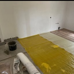 SZYMUR - Układanie paneli podłogowych w pokoju: folia paroizolacyjna, podkład, panele. Widoczne narzędzia i materiały budowlane. Etap prac wykończeniowych.
