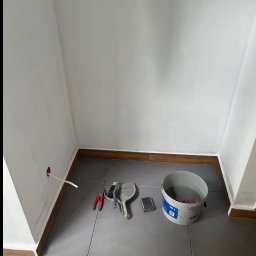 SZYMUR - Wnętrze w trakcie odświeżania: białe ściany z widocznym gniazdkiem, szare płytki podłogowe, wiadro z wodą i narzędzia malarskie. Listwa przypodłogowa w kolorze drewna.