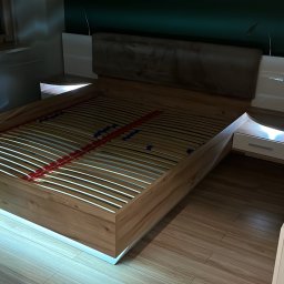 SZYMUR - Drewniane łóżko z podświetleniem LED i stelażem listwowym, widoczne z perspektywy pokoju, z elementami szafek nocnych i wezgłowia. Ustawione na panelach podłogowych.