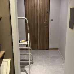 SZYMUR - Korytarz w trakcie odświeżania: drabina malarska, zabezpieczone taśmą krawędzie sufitu i drzwi. Widoczne płytki podłogowe i świeżo pomalowane ściany w odcieniach szarości.