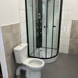 SZYMUR - Nowoczesna kabina prysznicowa z czarną ramą i biała toaleta w wykończonej łazience z szarymi płytkami podłogowymi i częściowo wyłożonymi ścianami.