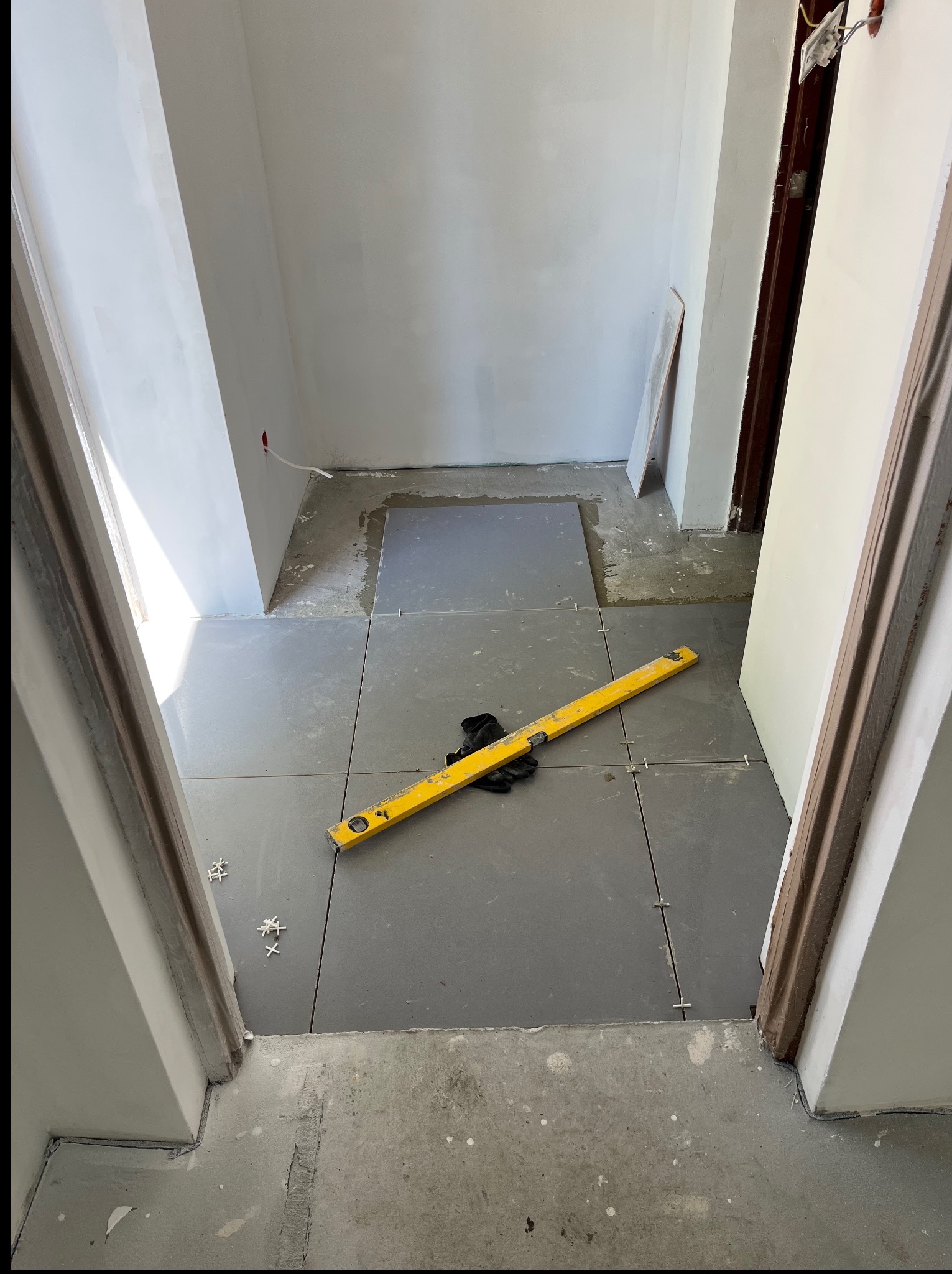 Szare płytki podłogowe ułożone w korytarzu, poziomica i rękawice robocze na powierzchni. Widok z perspektywy wejścia, fragment ościeżnicy.
