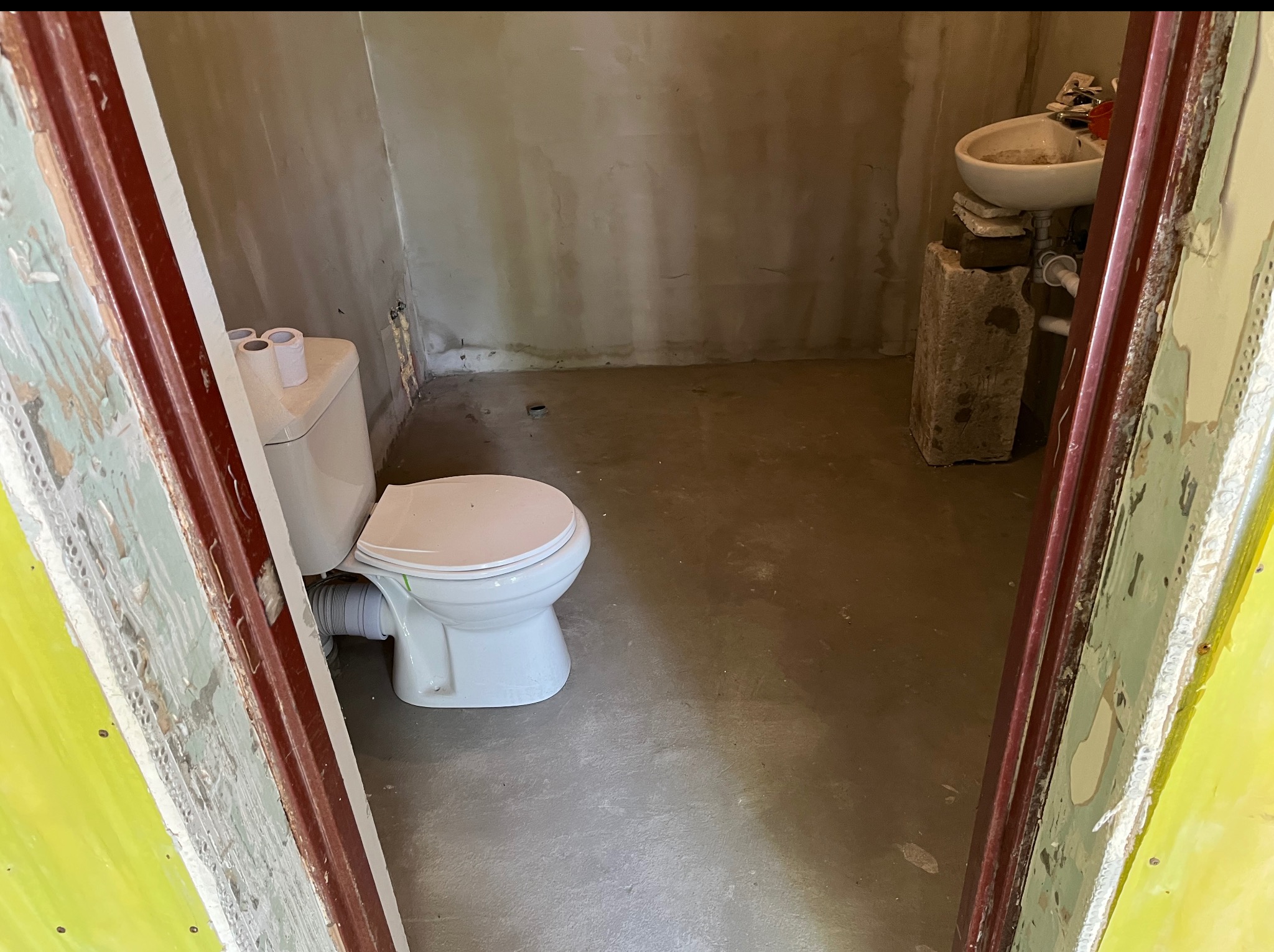 Surowe wnętrze łazienki w trakcie remontu: świeżo wylana posadzka, zamontowana toaleta, umywalka na prowizorycznym postumencie z cegieł, widoczne ślady po starych powłokach.