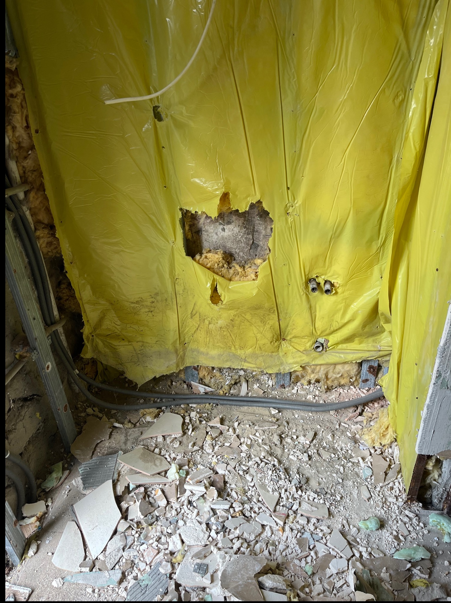 Fragment ściany w trakcie demontażu, zabezpieczonej żółtą folią, z widocznymi instalacjami i gruzem na podłodze. Widoczne przewody elektryczne i rury.
