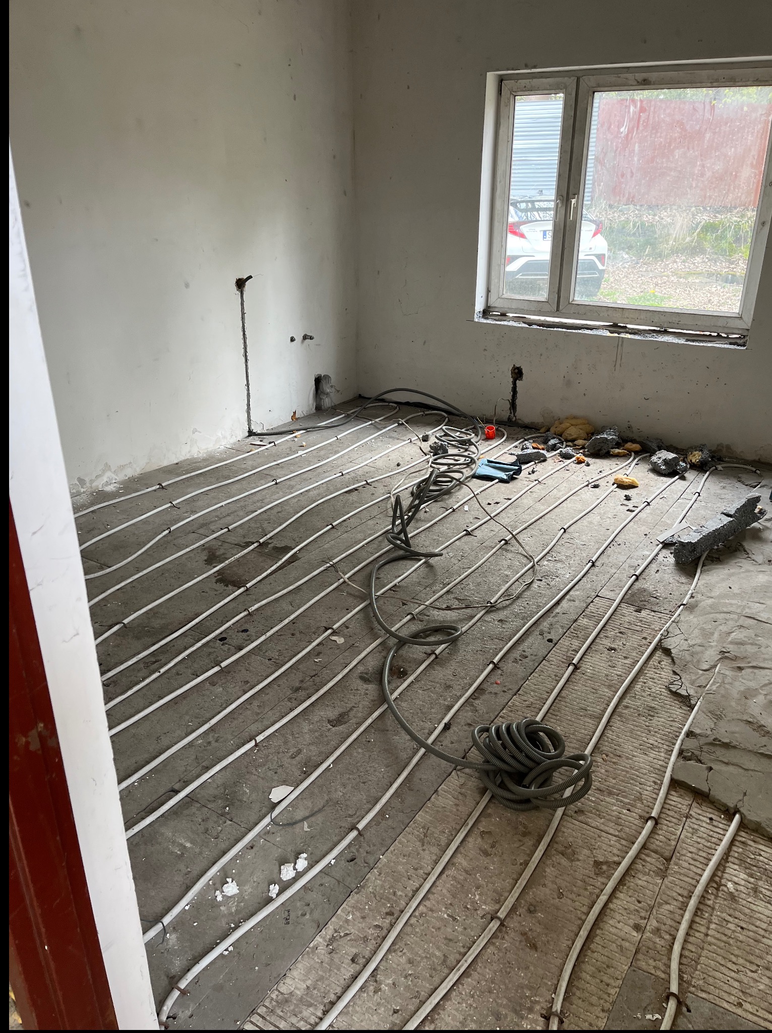 Białe rurki ogrzewania podłogowego ułożone równolegle na szarym, surowym podłożu w remontowanym pokoju z oknem. Widoczne kable i materiały budowlane.